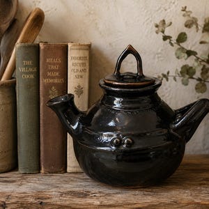 May include: A dark brown, glazed ceramic teapot with a rounded body, a short spout, and a pointed lid handle. The teapot features two small, raised circular decorative accents on the front. It is positioned next to three vintage books.