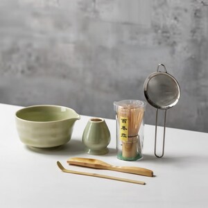 May include: A matcha tea set comprising a pale green ceramic bowl, a small vase, a bamboo whisk, a bamboo scoop, a metal sieve, and a container of bamboo whisk. The items are arranged on a white surface.