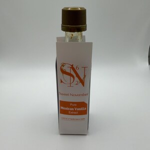 Op de afbeelding: Een kleine glazen fles Sweet November Pure Mexican Vanilla Extract. De fles heeft een bruine dop en is gedeeltelijk bedekt met een wit etiket met oranje tekst. Op het etiket staat "Sweet November" en "Pure Mexican Vanilla Extract".