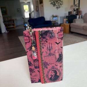 May include: A rectangular book-shaped box with a pink and black gothic-themed cover featuring illustrations of a castle, bats, a candelabra, and a portrait of a man. A silver-toned metal charm with a small glass vial containing pink glitter is attached.