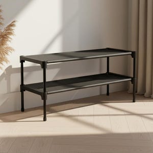 May include: A two-tiered, black metal shoe rack with a rectangular design. The rack is positioned against a white wall, with a vase of dried plants to the left. The room has a neutral color palette with natural light.