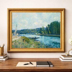 May include: A framed landscape painting with a gold ornate frame, depicting a river scene with a sailboat and trees. The painting is displayed on a wooden desk with a lamp, books, and a vase of white flowers.