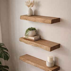 Pode incluir: Três prateleiras flutuantes de madeira montadas numa parede branca. As prateleiras são feitas de madeira castanha clara e têm uma borda natural. A prateleira superior contém um vaso branco com flores secas. A prateleira do meio tem uma pequena planta em vaso e livros. A prateleira inferior tem uma vela.