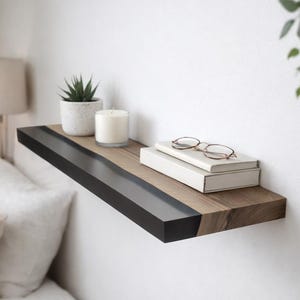 May include: A wall-mounted floating shelf featuring a natural wood grain surface with a contrasting matte black front edge. The shelf holds a small potted succulent, a white pillar candle, two stacked books, and a pair of thin-rimmed glasses.