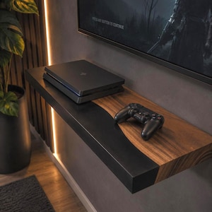 May include: A wall-mounted floating shelf featuring a combination of natural brown wood grain and a solid matte black section. A black gaming console and a black wireless game controller are placed on the surface of the shelf below a television.