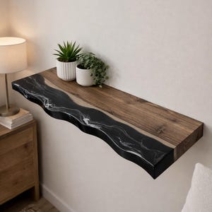 May include: A floating shelf with a live edge design. The shelf features a dark wood grain and a black epoxy resin with white marbling. Two small potted plants sit on top of the shelf.