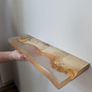 Op de afbeelding: Een houten plank met een natuurlijke rand en een transparante hars voorkant. Het hout heeft een natuurlijke nerf met bruine en bruinachtige tinten. De hars heeft een doorschijnende amberkleur. De plank is aan een witte muur bevestigd.