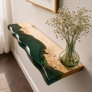 Puede incluir: Un estante flotante de pared que presenta una losa de madera clara natural con borde irregular y una incrustación de resina verde oscuro translúcida que imita un río. Un florero de vidrio transparente con flores blancas descansa sobre él.