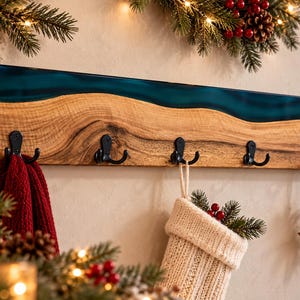 May include: A rectangular wall-mounted coat rack featuring a natural wood grain board with a wavy teal resin inlay along the top edge. The rack includes five matte black metal hooks, with a red towel, a knit stocking, and a wooden star ornament.