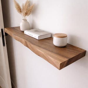 Könnte beinhalten: Ein schwebendes Holzregal mit roher Kante, an einer weißen Wand befestigt. Das Regal enthält eine weiße Vase mit getrocknetem Pampasgras, ein Buch mit dem Wort "KINfolk" auf dem Einband und ein kleines weißes Glas mit Holzdeckel. Das Regal ist aus dunkelbraunem Holz.
