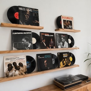 May include: Three horizontal light brown wooden floating shelves mounted on a wall, displaying various vinyl record albums and black discs. The collection includes titles like Miles Davis Kind of Blue, John Coltrane A Love Supreme, and Thelonious Monk.