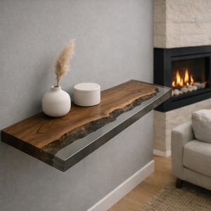 May include: A wall-mounted wooden shelf with a live edge, showcasing a dark wood grain and a clear resin inlay. A white vase with dried pampas grass and a small white container are on the shelf. The shelf is fixed to a grey wall.