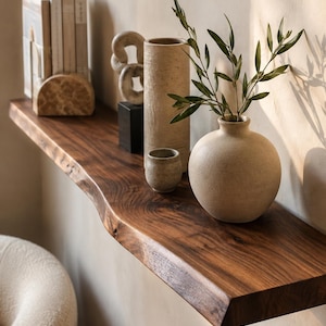 Op de afbeelding: Een zwevende wandplank van donker hout met een natuurlijke rand, waarop decoratieve voorwerpen staan, waaronder een ronde beige keramische vaas met groene bladeren, een klein bijpassend kopje, een hoge cilindrische vaas, een stenen sculptuur en boeken.
