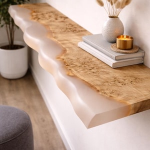 May include: A wooden floating shelf with a live edge design. The shelf has a light-coloured resin edge and natural wood grain. A white vase with dried flowers, books, and a candle sit on the shelf. The shelf is mounted on a white wall.
