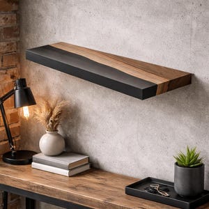 May include: A wall-mounted wooden shelf with a black trim and a natural wood grain finish. The shelf is fixed to a grey concrete wall. A black desk lamp, books, and a small plant are on a wooden table below.