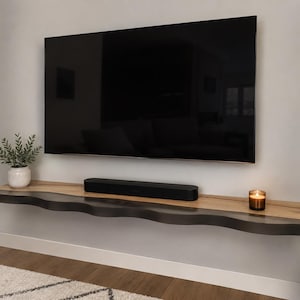 Peut inclure: Une étagère murale flottante avec une surface supérieure en bois clair naturel et un bord avant sombre et ondulé. L'étagère supporte une barre de son noire, une petite plante en pot blanche et un bougeoir en verre sous un grand téléviseur.