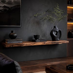 Peut inclure: Une étagère flottante en bois à bord brut supportant un vase sphérique noir, une pile de livres intitulée The Kinfolk, un petit bol noir sur des livres et un grand vase sculptural noir avec une ouverture centrale contenant des branches feuillues.