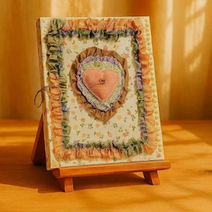 May include: A decorative, handmade heart-shaped wall hanging on a small wooden easel. The heart is made of pink fabric and surrounded by layers of ruffled fabric in shades of brown, green, and purple. The background is a floral pattern.