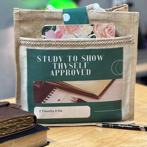 May include: A tan burlap tote bag featuring a front pocket with a green rectangular graphic. The graphic displays the text "STUDY TO SHOW THYSELF APPROVED" and "2 Timothy 2:15a" above an illustration of a notebook and pen. A floral notebook is tucked inside.