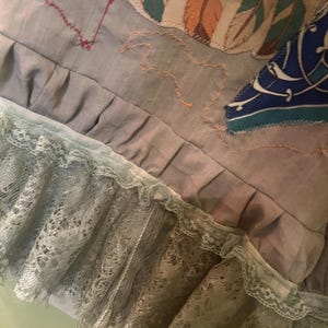 May include: A grey fabric garment featuring a ruffled hem with layered light green lace trim. The surface displays embroidered feather motifs in teal and orange, a red stitched rectangular outline, and a blue patterned fabric patch.