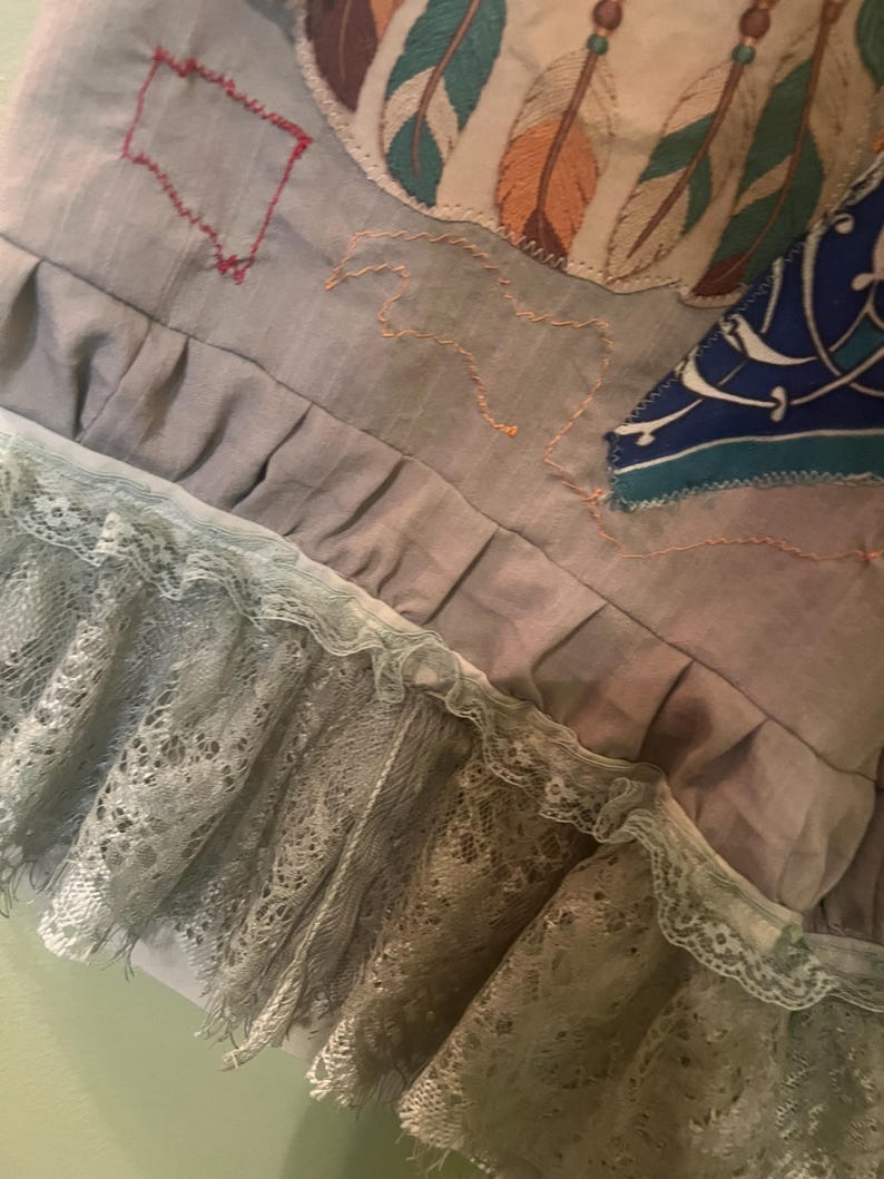 May include: A grey fabric garment featuring a ruffled hem with layered light green lace trim. The surface displays embroidered feather motifs in teal and orange, a red stitched rectangular outline, and a blue patterned fabric patch.