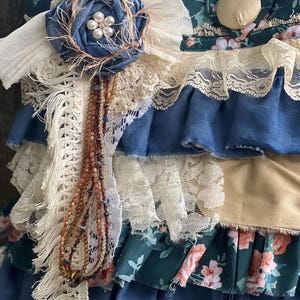 Peut inclure: Gros plan sur un article en tissu fait &agrave; la main avec des couches de tissus bleus, cr&egrave;me et &agrave; motifs floraux. Une fleur en tissu bleu avec un centre de perles et un brin de perles ajoutent des d&eacute;tails d&eacute;coratifs. L'article a une esth&eacute;tique vintage et boh&egrave;me.