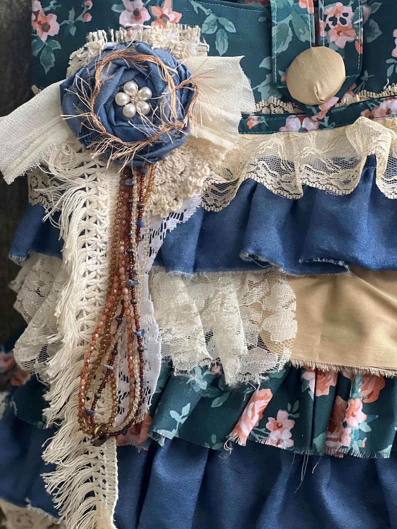 Peut inclure: Gros plan sur un article en tissu fait &agrave; la main avec des couches de tissus bleus, cr&egrave;me et &agrave; motifs floraux. Une fleur en tissu bleu avec un centre de perles et un brin de perles ajoutent des d&eacute;tails d&eacute;coratifs. L'article a une esth&eacute;tique vintage et boh&egrave;me.
