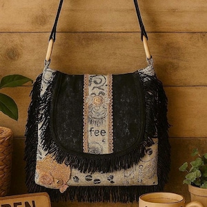 May include: A black and beige shoulder bag with a flap closure, fringe trim, and wooden handles. The bag features a decorative design with the word "fee" on the front. A coffee cup with the word "BEAN" is also visible.