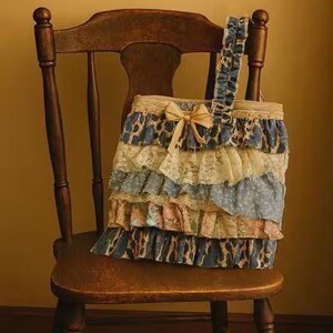 Peut inclure: Un sac fourre-tout d&eacute;coratif avec des volants superpos&eacute;s bleus, cr&egrave;me et roses, orn&eacute; d'un n&oelig;ud beige. Le sac est pos&eacute; sur une chaise en bois vintage.