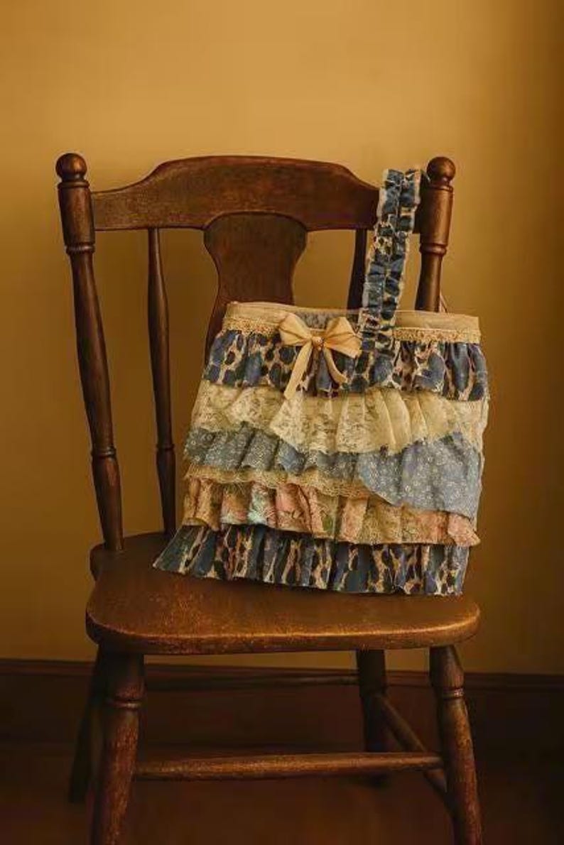 Peut inclure: Un sac fourre-tout d&eacute;coratif avec des volants superpos&eacute;s bleus, cr&egrave;me et roses, orn&eacute; d'un n&oelig;ud beige. Le sac est pos&eacute; sur une chaise en bois vintage.