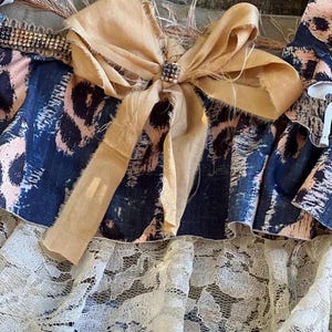 Peut inclure: Gros plan sur un textile d&eacute;coratif avec un grand n&oelig;ud beige orn&eacute; d'un carr&eacute; de strass. Le textile pr&eacute;sente un imprim&eacute; animalier bleu et rose, une bordure perl&eacute;e et une couche de dentelle blanche.
