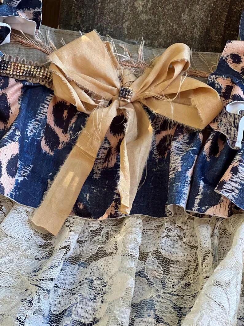 Peut inclure: Gros plan sur un textile d&eacute;coratif avec un grand n&oelig;ud beige orn&eacute; d'un carr&eacute; de strass. Le textile pr&eacute;sente un imprim&eacute; animalier bleu et rose, une bordure perl&eacute;e et une couche de dentelle blanche.
