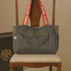 May include: Olive green tote bag with red straps that read "REMOVE BEFORE FLIGHT." The bag has multiple pockets and a military-inspired design. It is displayed on a wooden chair with a cream-colored seat. A dark green box is visible in the background.