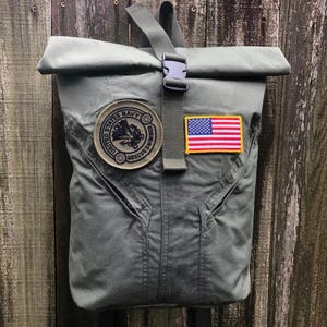 May include: Olive green backpack with a roll-top closure and a black buckle. The bag features a patch with the words "United States Navy Rescue Swimmer" and an American flag patch.