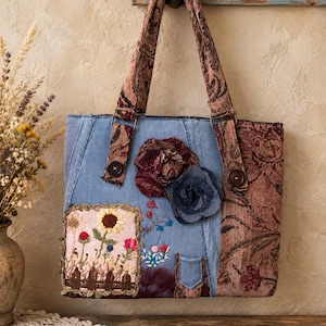 May include: A tote bag constructed from light blue denim and dark floral tapestry fabric. The front features a rectangular patch with embroidered sunflowers and flowers, two fabric flower appliqués, and small embroidered floral details.