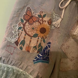 May include: Light grey shorts featuring a drawstring waist, a ruffled lace hem, and a large embroidered patch depicting a dreamcatcher with a sunflower, a monarch butterfly, and hanging feathers, accented by additional fabric patches and lace trim.