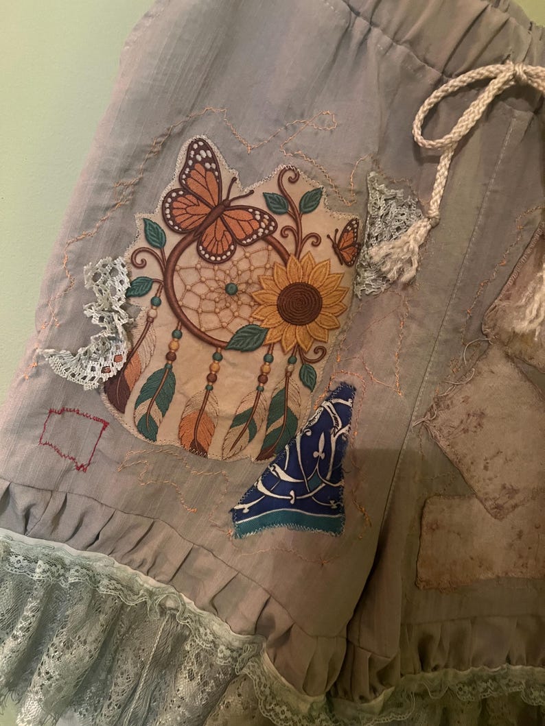 May include: Light grey shorts featuring a drawstring waist, a ruffled lace hem, and a large embroidered patch depicting a dreamcatcher with a sunflower, a monarch butterfly, and hanging feathers, accented by additional fabric patches and lace trim.