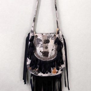 May include: A white and black patterned crossbody bag with black fringe and ribbon accents. The bag features decorative buttons and a silver-toned trim. The strap is patterned to match the bag's design.