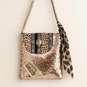 May include: A rectangular shoulder bag with a sequined front and a leopard print flap. The bag features a braided rope strap and a decorative leopard print scarf. A gold-colored metal clasp secures the flap.