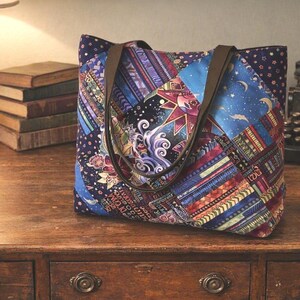 May include: A tote bag featuring a patchwork design with various patterns including celestial suns, moons, stars, and abstract geometric shapes in shades of blue, purple, and gold. The bag has two brown fabric shoulder straps and a rectangular shape.