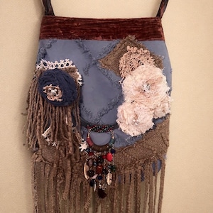 Puede incluir: Bolso de hombro artesanal con base azul similar al ante, adornado con parches texturizados, flecos y elementos decorativos. Cuenta con una parte superior de terciopelo marrón oscuro y un colgante de cuentas con acentos coloridos. La correa es marrón oscuro.