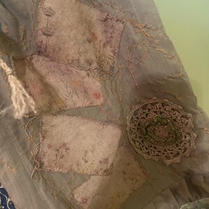 May include: A light grey fabric garment featuring rectangular patches with faded floral prints, accented by orange decorative stitching. A circular, textured, light brown crocheted applique is attached to the side, and a light grey lace trim borders the bottom edge.