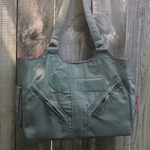 Puede incluir: Bolso tote verde oliva con un diseño único. Cuenta con un parche circular, un parche rectangular y dos bolsillos con cremallera. El bolso tiene una correa para el hombro y un forro estampado. El bolso está hecho de un material duradero.