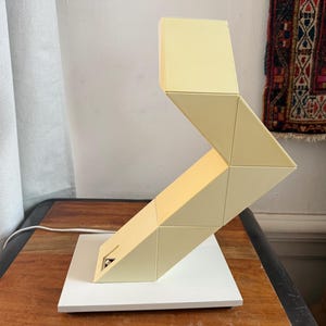 Op de afbeelding: Een moderne, geometrische bureaulamp in crèmekleur. De lamp heeft een reeks onderling verbonden driehoekige en rechthoekige vormen, waardoor een zigzagontwerp ontstaat. Hij staat op een witte vierkante basis en heeft een wit netsnoer. De lamp staat op een houten tafel.