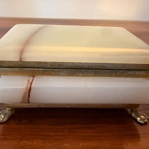 Può includere: Un portagioie rettangolare con coperchio incernierato. La scatola è realizzata in onice bianco e crema con una venatura marrone-rossastra che attraversa la pietra. La scatola ha dettagli in ottone e poggia su quattro piedini a zampa d'ottone.