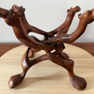 May include: A handcrafted wooden sculpture featuring three stylized camel figures intertwined to form a decorative stand. The camels are a rich brown color, with detailed carving. The stand is placed on a light-colored wooden base.