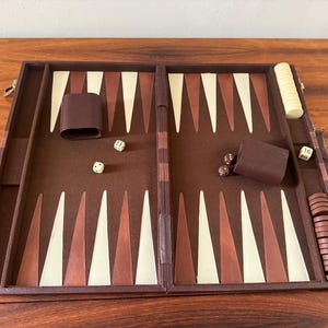 May include: A brown backgammon set with a dark wood exterior. The game board is open, revealing brown and cream-colored triangles. Dice, dice cups, and game pieces are visible. The set is displayed on a dark wood surface.