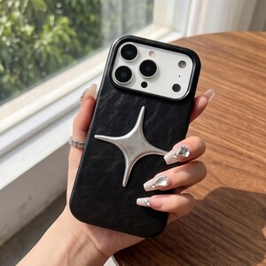 Puede incluir: Una funda de smartphone negra con acabado de textura similar al cuero y un prominente emblema plateado en forma de estrella de cuatro puntas centrado en la parte posterior. La funda presenta un recorte elevado para la cámara con aberturas.