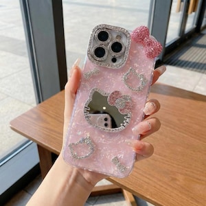 May include: Pink phone case with a glittery finish, featuring a Hello Kitty design with a mirror face and a bow. The case is adorned with rhinestones around the camera and the Hello Kitty design, and a pink bow at the top.