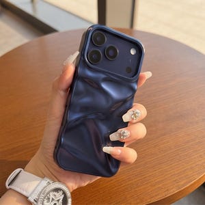 Puede incluir: Una funda para smartphone de color azul marino oscuro con una textura superficial distintivamente ondulada, irregular y arrugada. La funda presenta un recorte rectangular elevado para la cámara con tres lentes circulares y un flash.